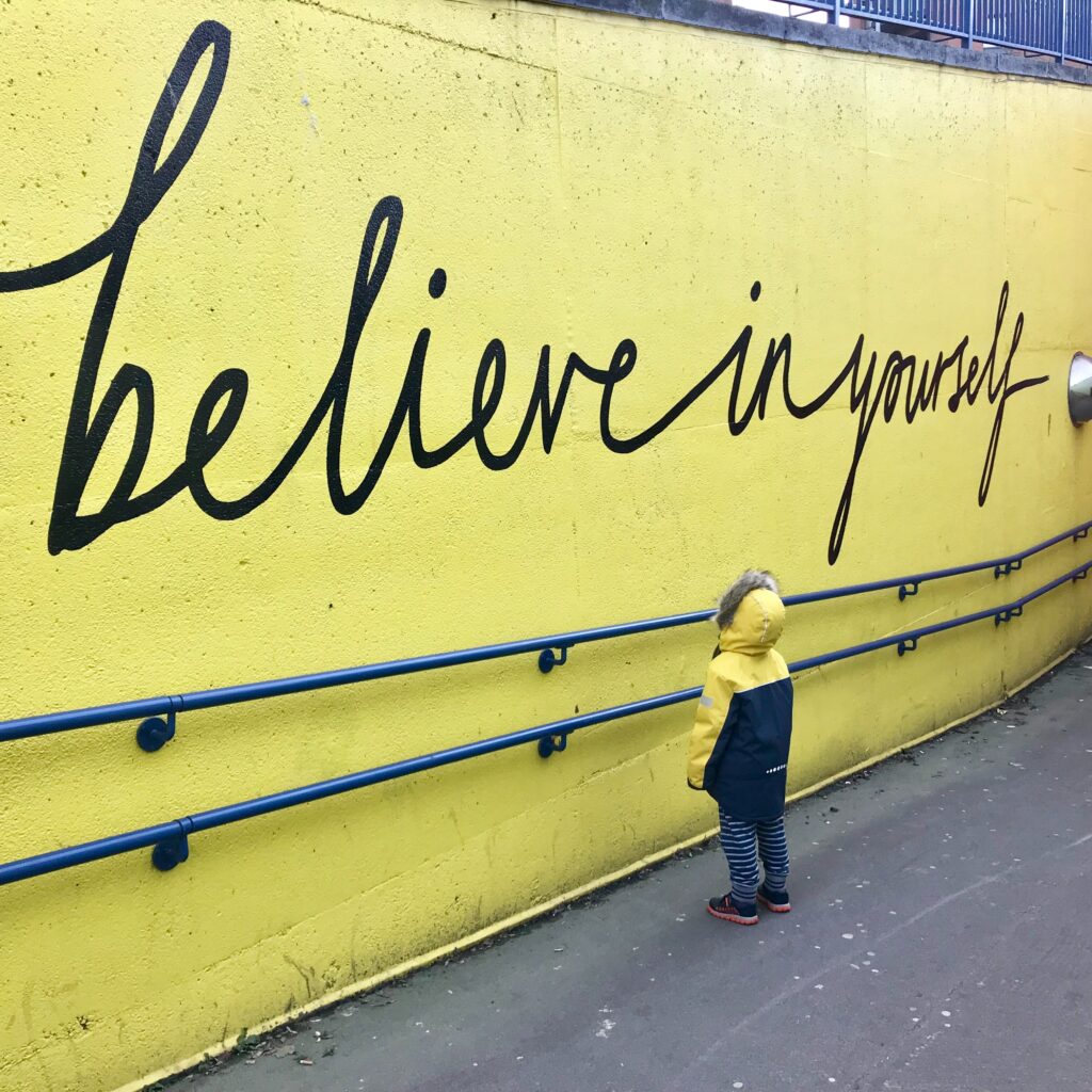 A child looking upon a mural that reads "believe in yourself" by Katrina Wright on Unsplash 